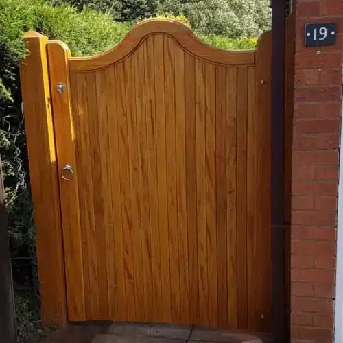 Link to our range of side gates showing a swan neck wooden side made from idigbo fitted to the side of a house and an oak post fitted into the bush at the property line. This side gate also has a lock and gate latch.